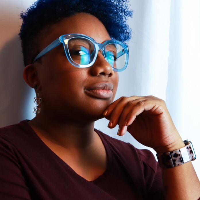 Black woman with blue hair and glasses smirks at the camera with hand on chin